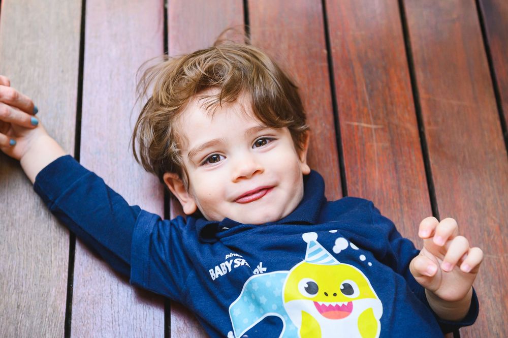 Young child in blue and yellow shark shirt lying on wooden deck while making peace sign gesture and smiling.
