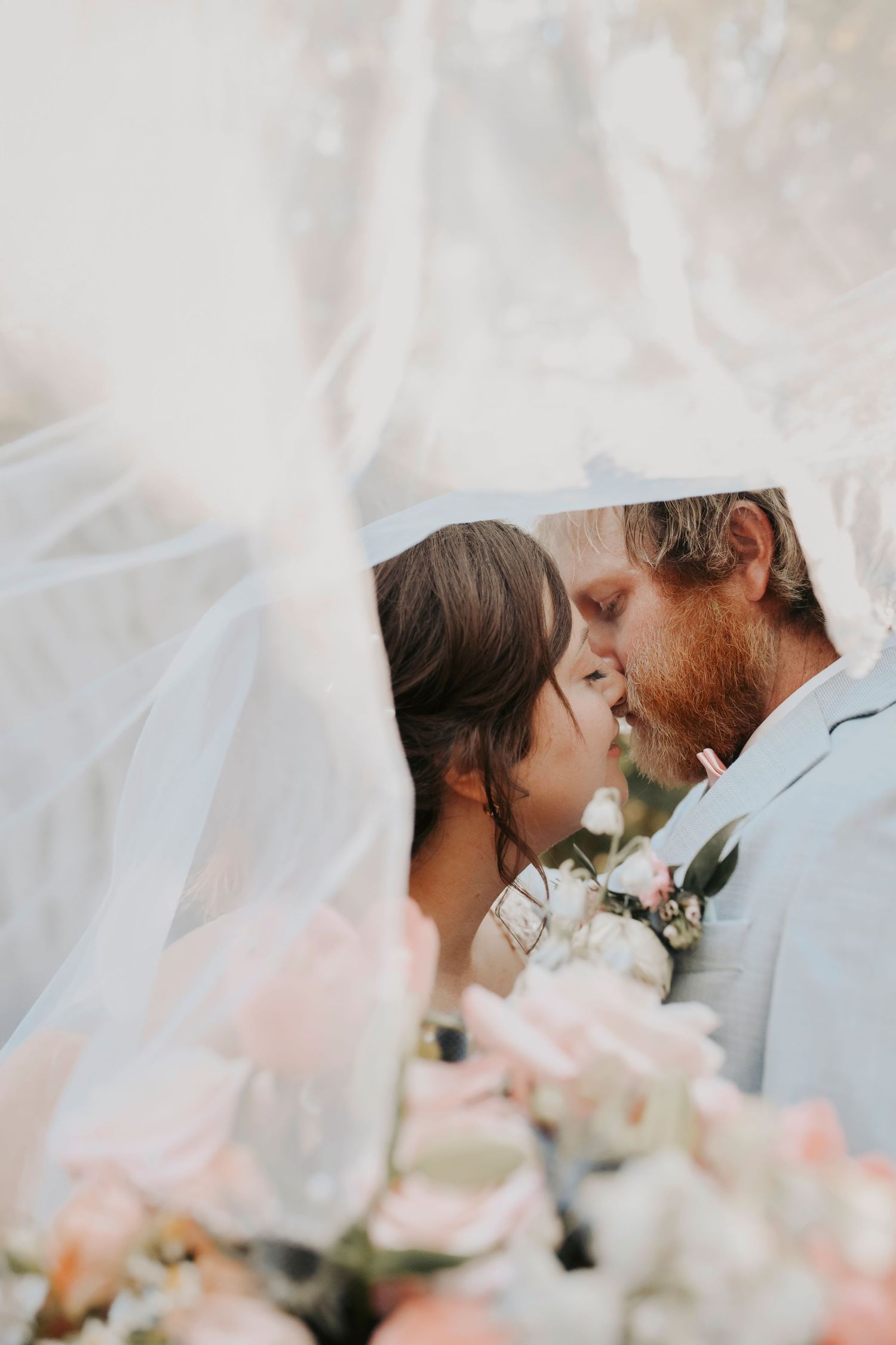 Bride and groom surrounded by pastel florals and fresh greenery kissing beneath her veil