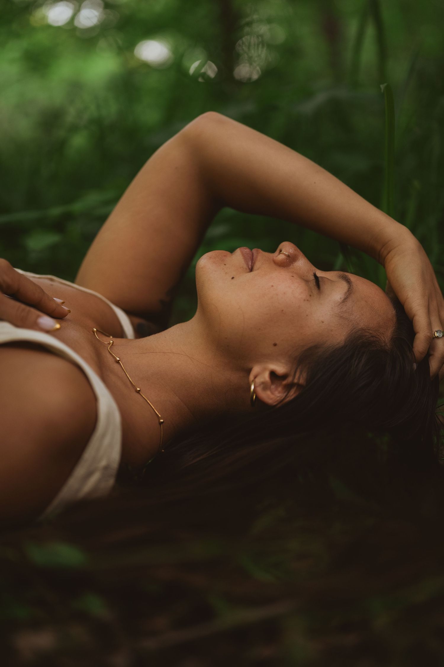 A person lies gracefully on soft ground in dappled sunlight among lush green foliage in a forest setting.