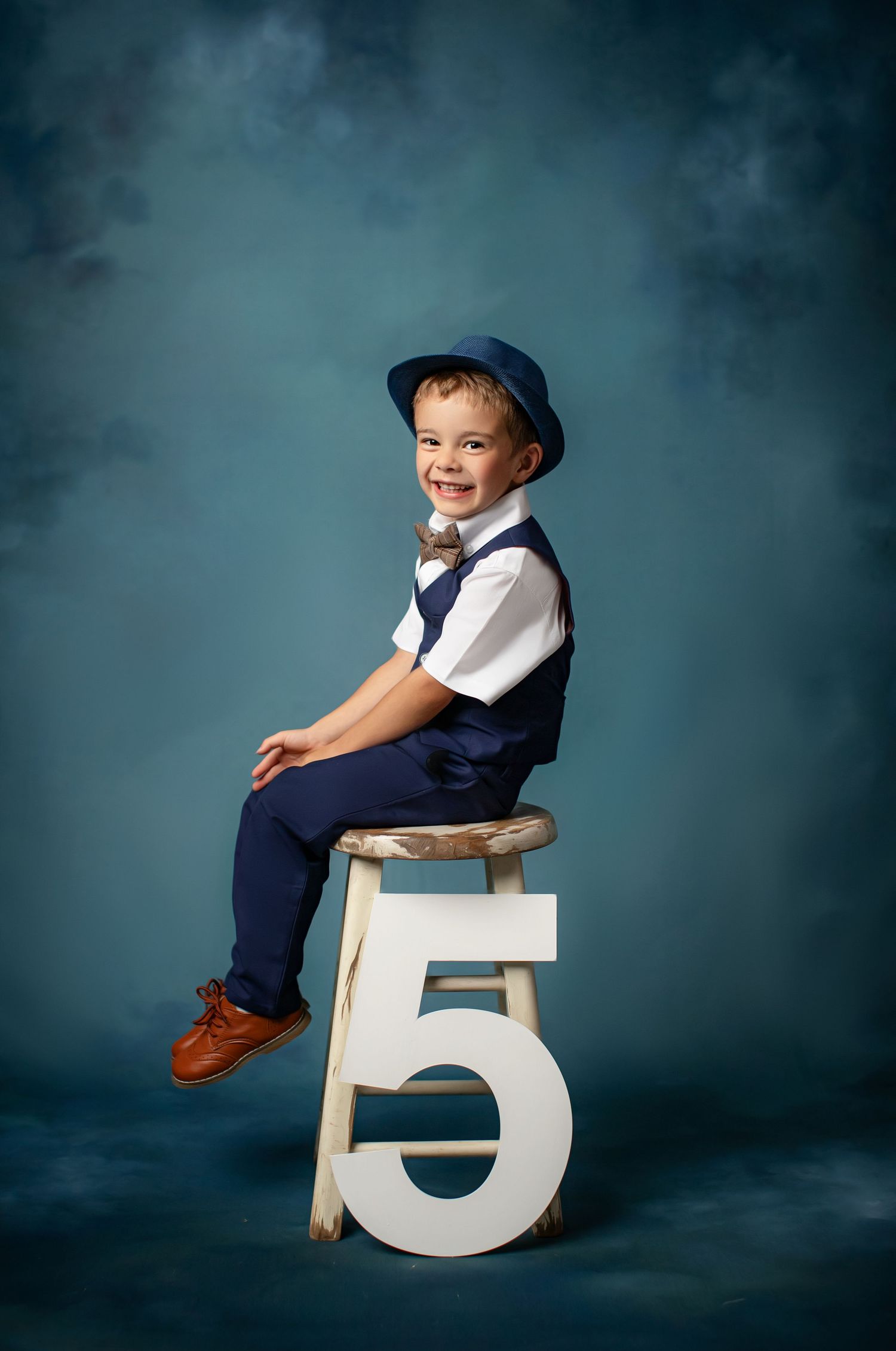 A young child in formal attire sits perched on a large decorative number 5 against a moody blue studio backdrop.