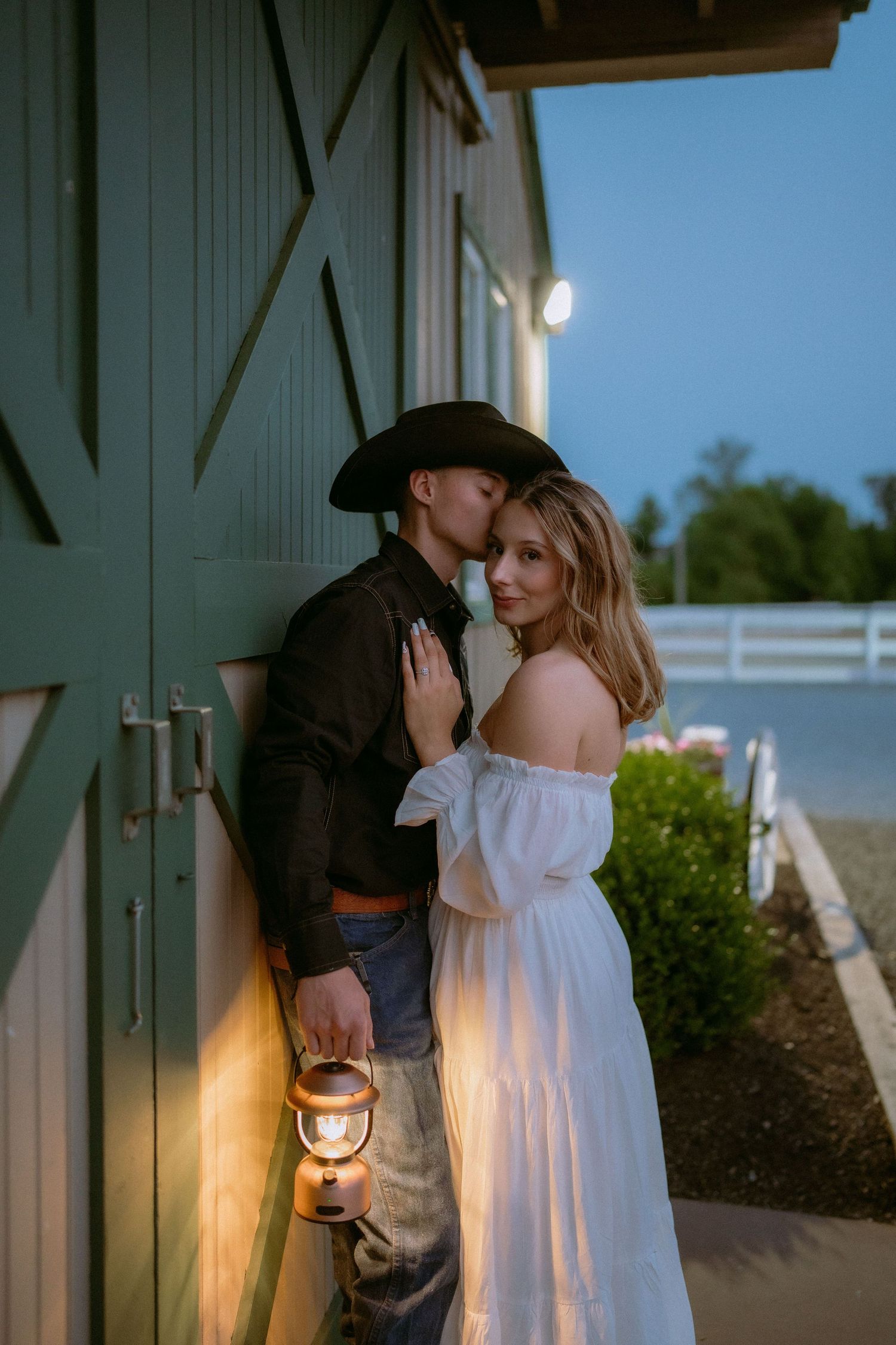 A romantic couple embraces by a rustic barn door at dusk with a cowboy holding a lantern.
