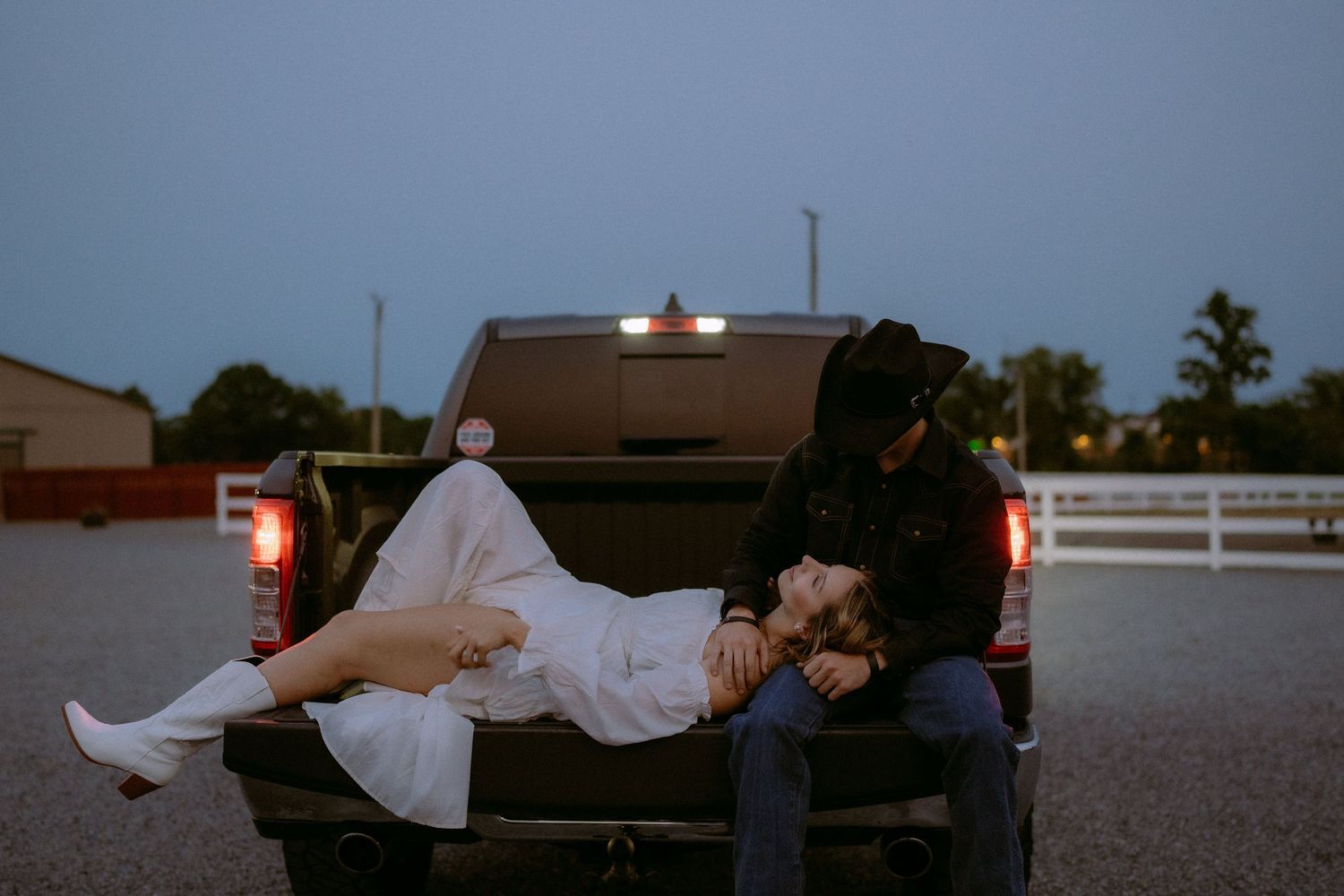 A couple in western attire poses on the back of a pickup truck at twilight.