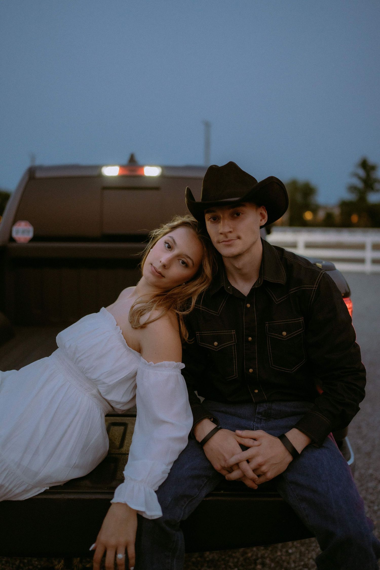A couple in western wear sits together in a pickup truck bed at sunset.