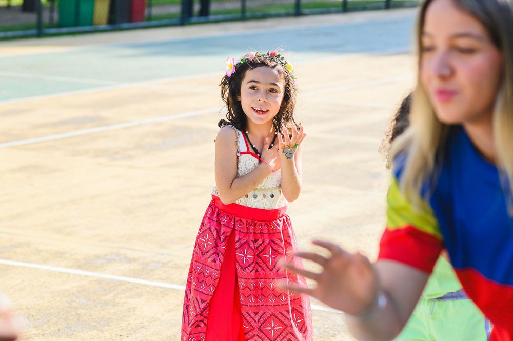 A young child in a bright pink skirt claps joyfully outdoors while another person stands nearby in blue clothing.