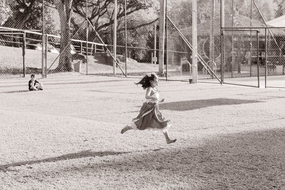 Vintage black and white photo of someone running joyfully in a dress across a playground with swings in the background.