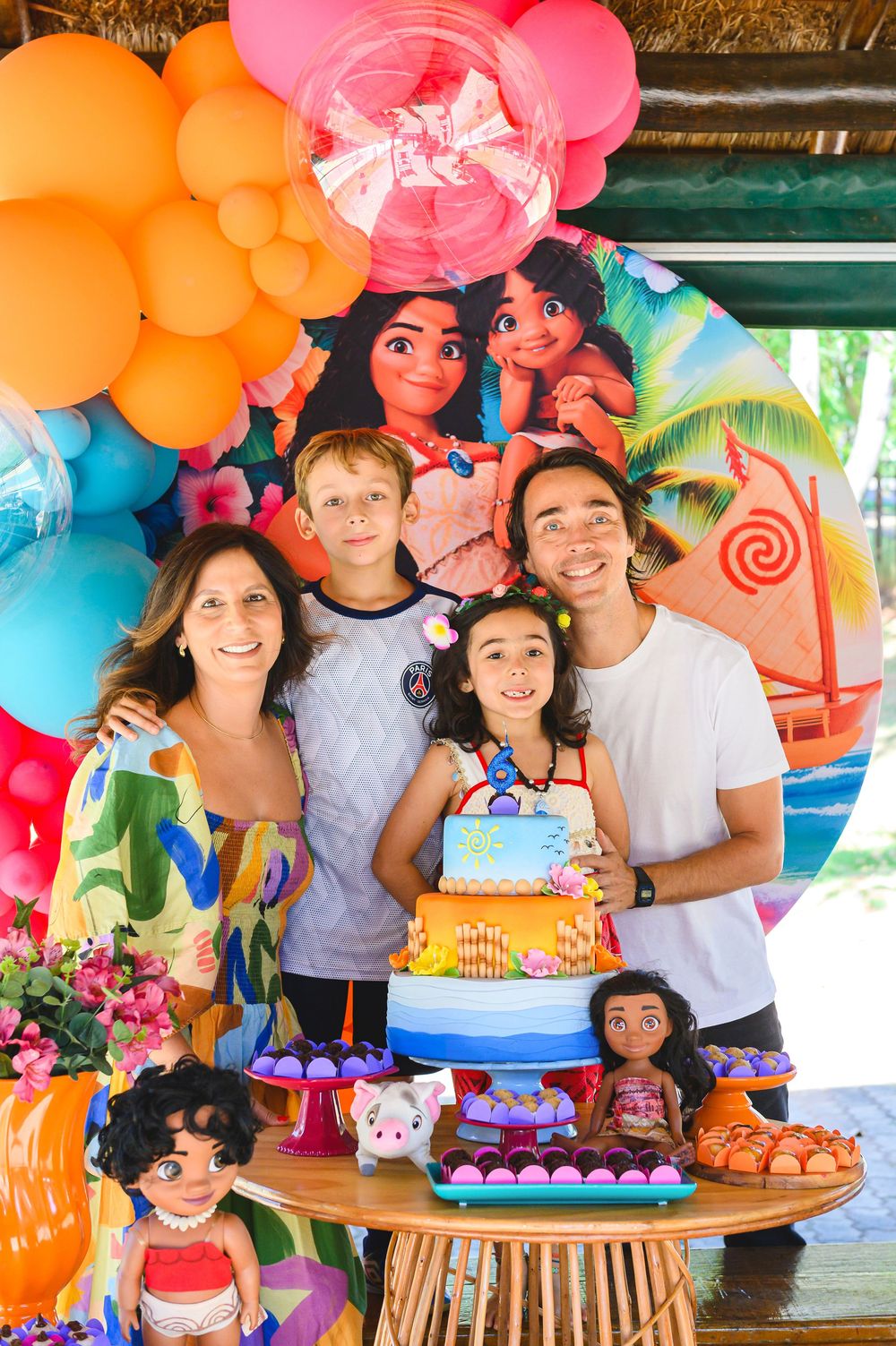 Colorful birthday party setup with bright balloon arch, Moana-themed decorations, and birthday cake on wooden table.