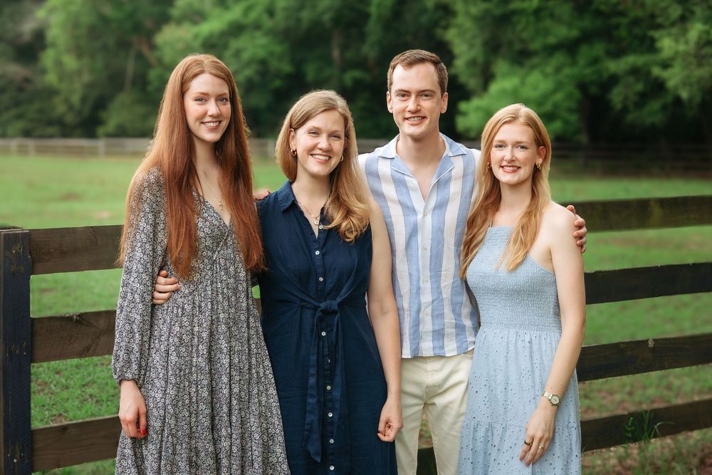 Four adult siblings smile together next to a fence during a family session in Athens, GA.