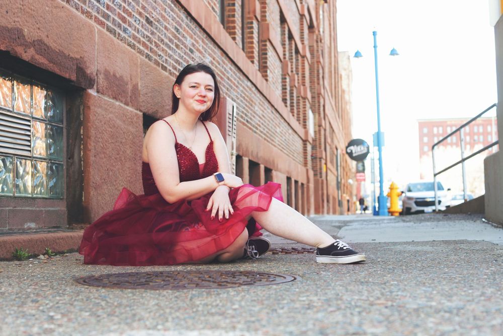 A person in a burgundy formal dress sits on a city sidewalk against a brick building on an urban street corner.