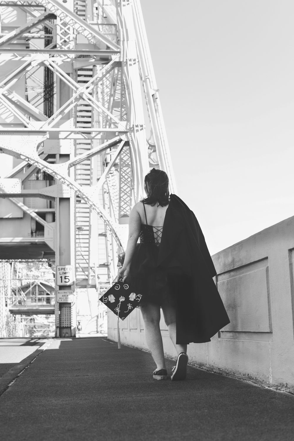 Black and white photo of a figure in a flowing cape walking past industrial bridge architecture.