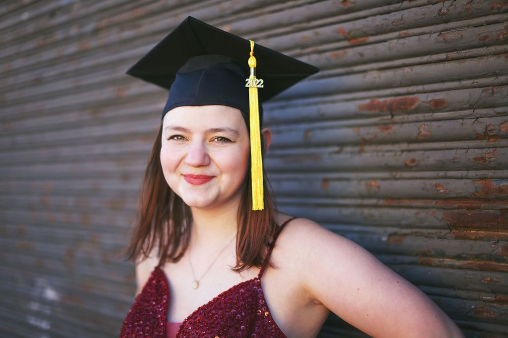 Graduate wearing a black cap with yellow tassel and burgundy sequined dress poses in front of weathered metal wall.
