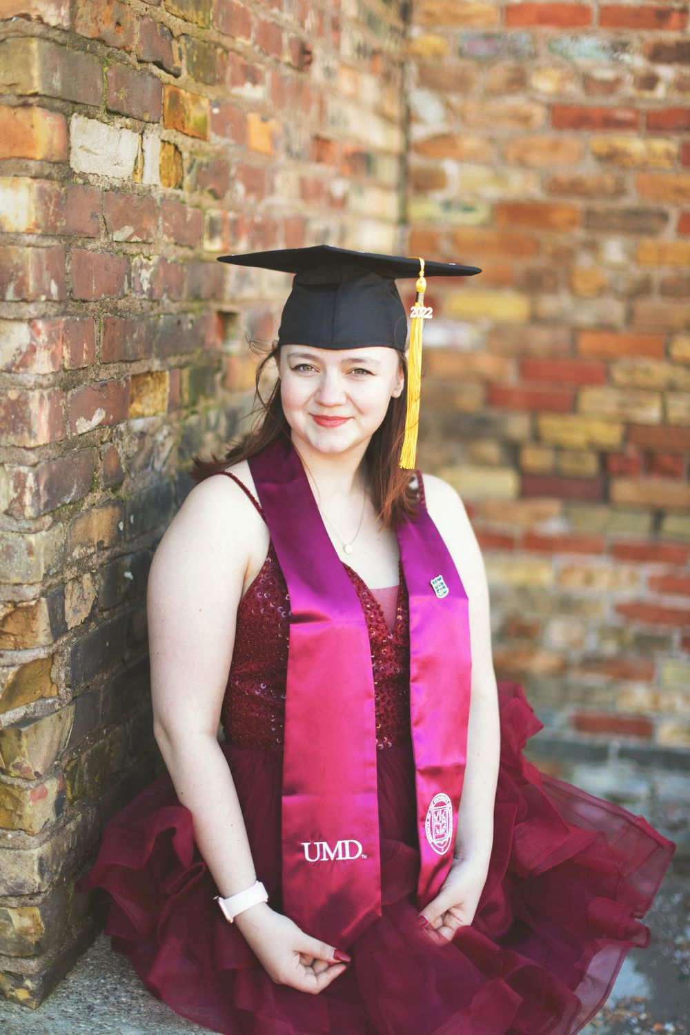 Graduation photos against a brick wall showing a graduate in burgundy dress and pink stole wearing academic regalia.