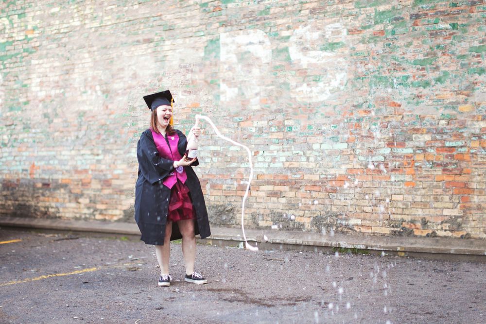 A graduate in cap and gown sprays champagne while celebrating against a brick wall background.