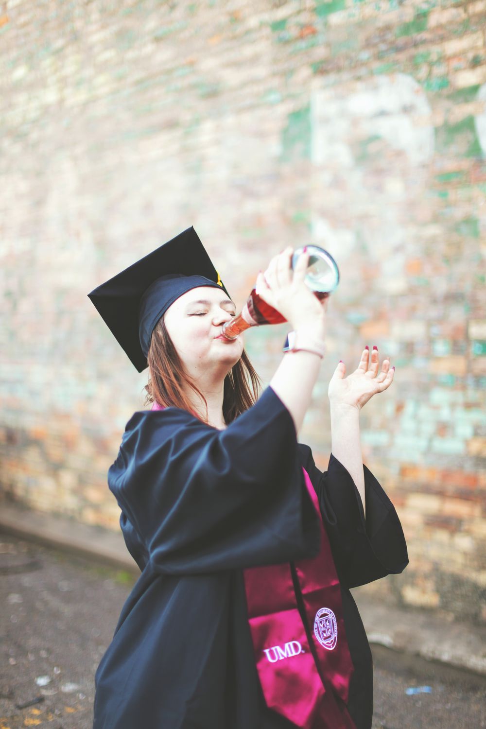 Graduate in cap and gown celebrates with diploma scroll and joyful pose against brick wall background.
