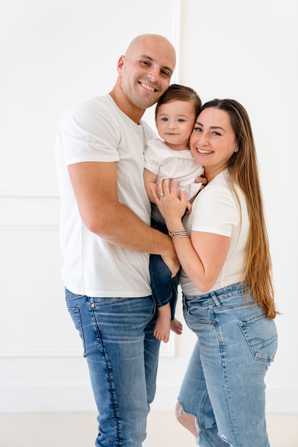 A couple in white t-shirts and jeans embrace their young child against a white background in a relaxed portrait.