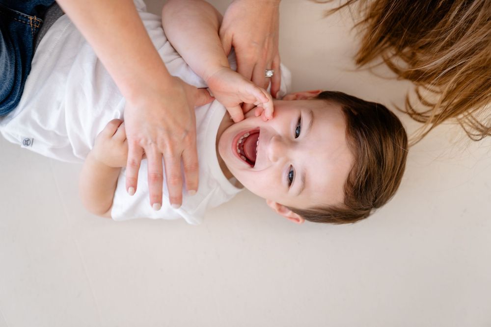 A smiling infant wearing a white onesie lies comfortably on a plush white surface in bright lighting.