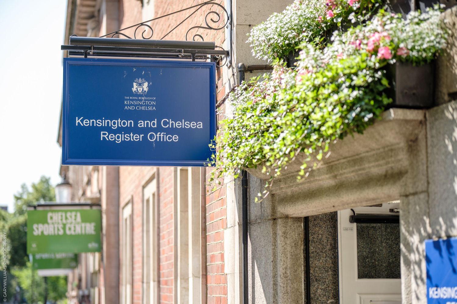 Blue sign for Registry Office hanging from brick building with white flowers cascading from above.
