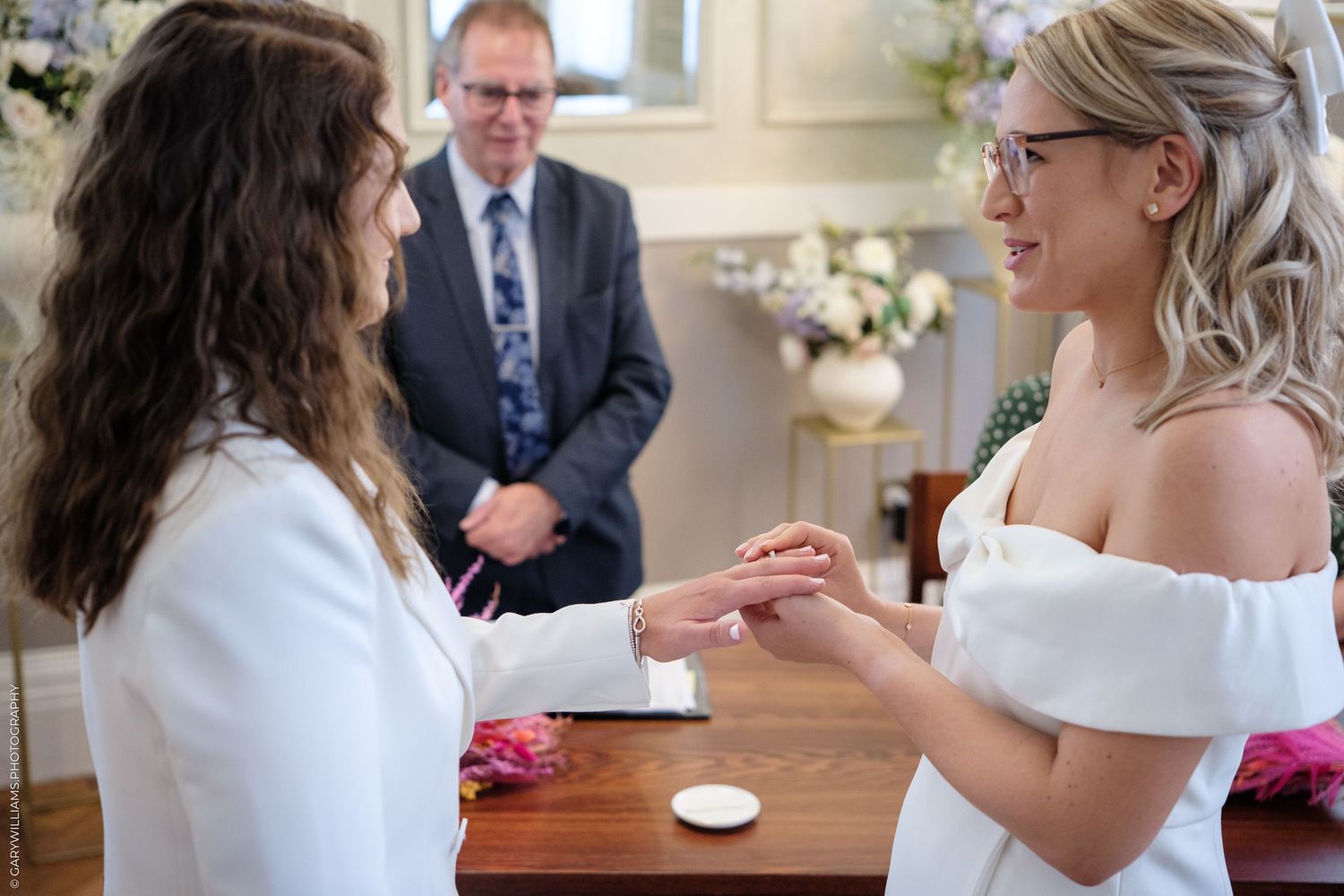 Two brides exchange rings during an intimate wedding ceremony while an officiant observes in the background.