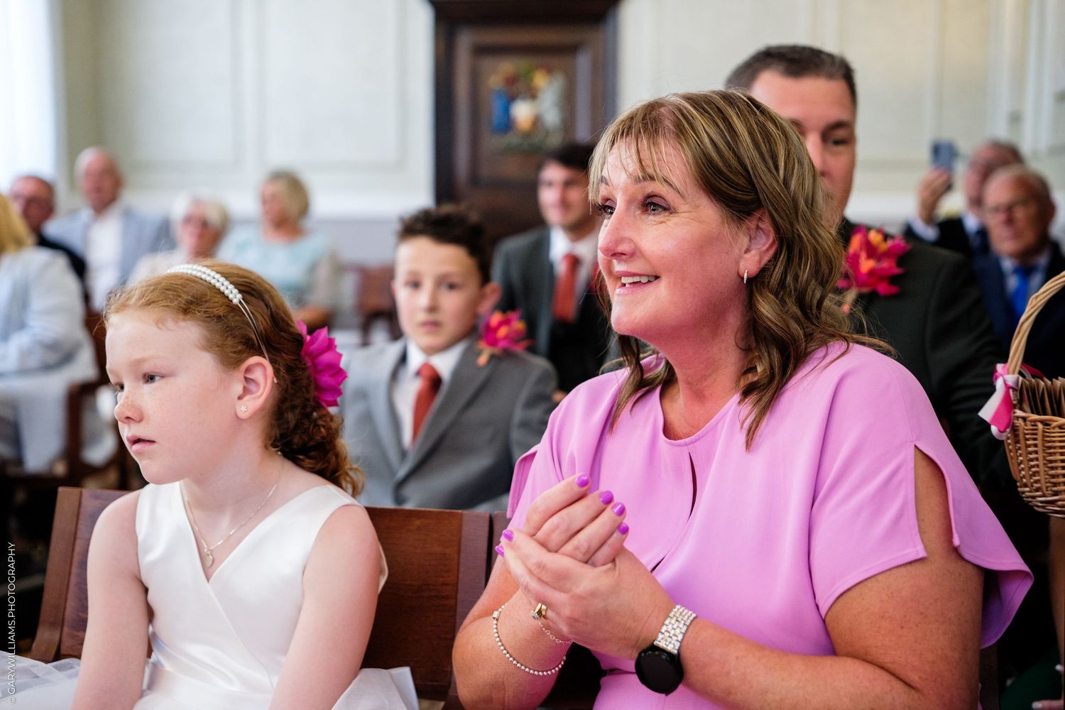 Two people sit in a church pew during a ceremony, with one wearing a white dress and another in pink attire.