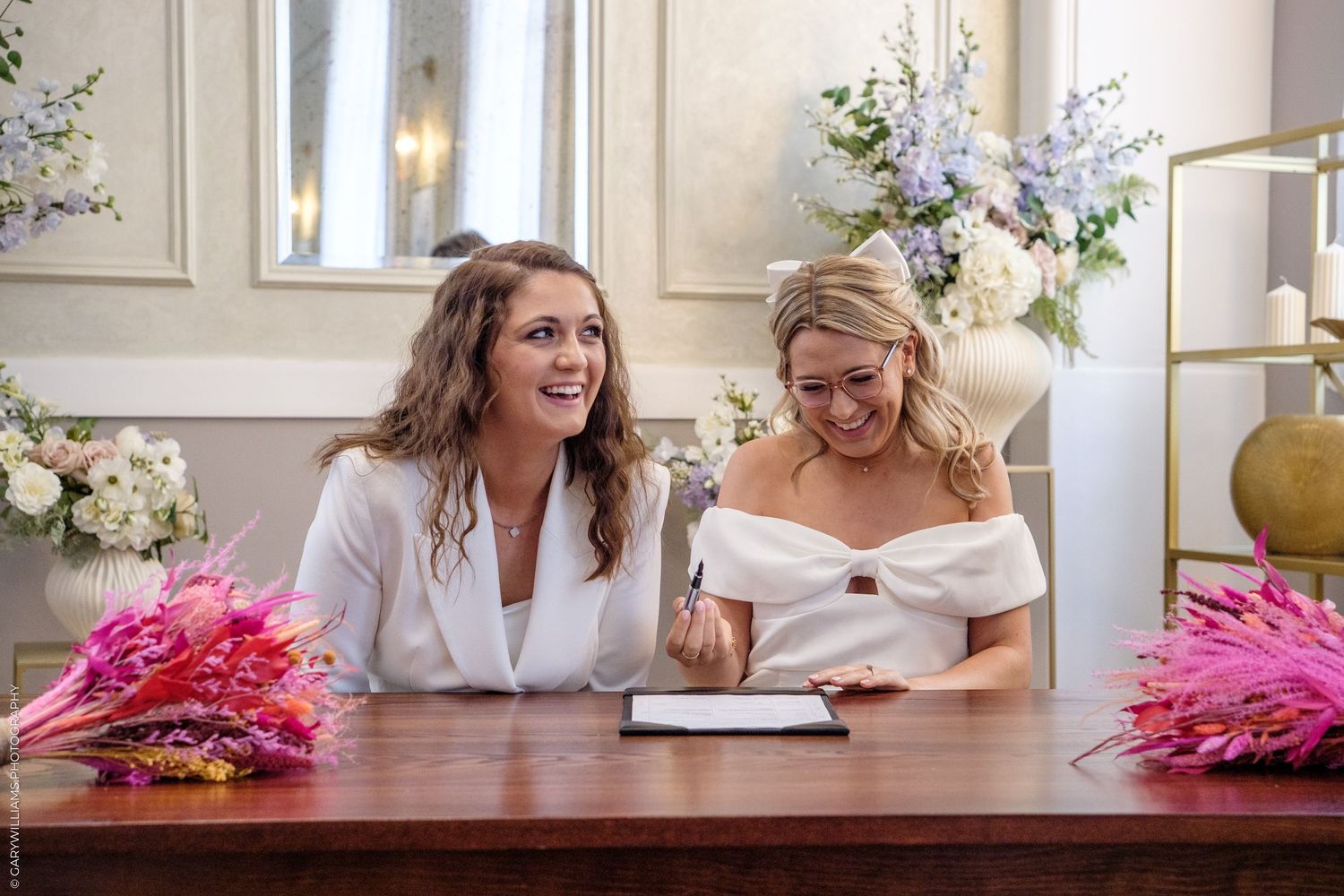 Two women sit around table admiring beautiful flower arrangements in wedding setting with soft natural lighting.
