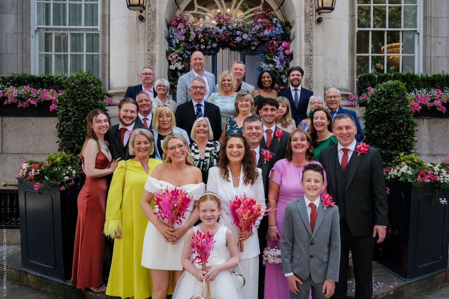 Wedding party gathers for formal photographs outside elegant venue with pink floral decorations.