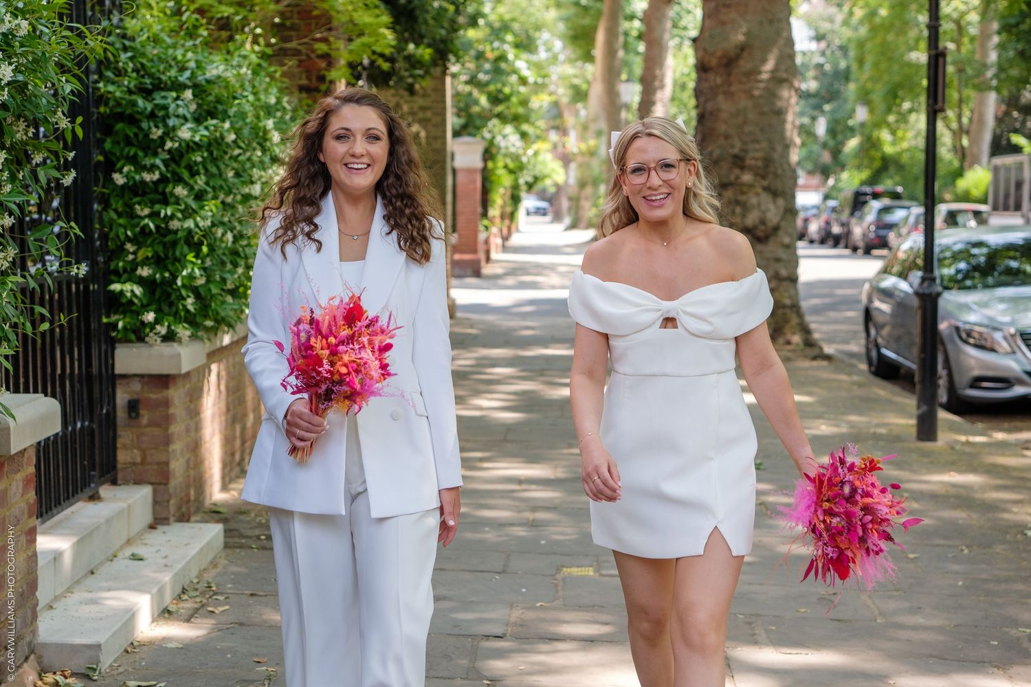 Two people in white outfits holding bright pink bouquets pose together on a tree-lined sidewalk.
