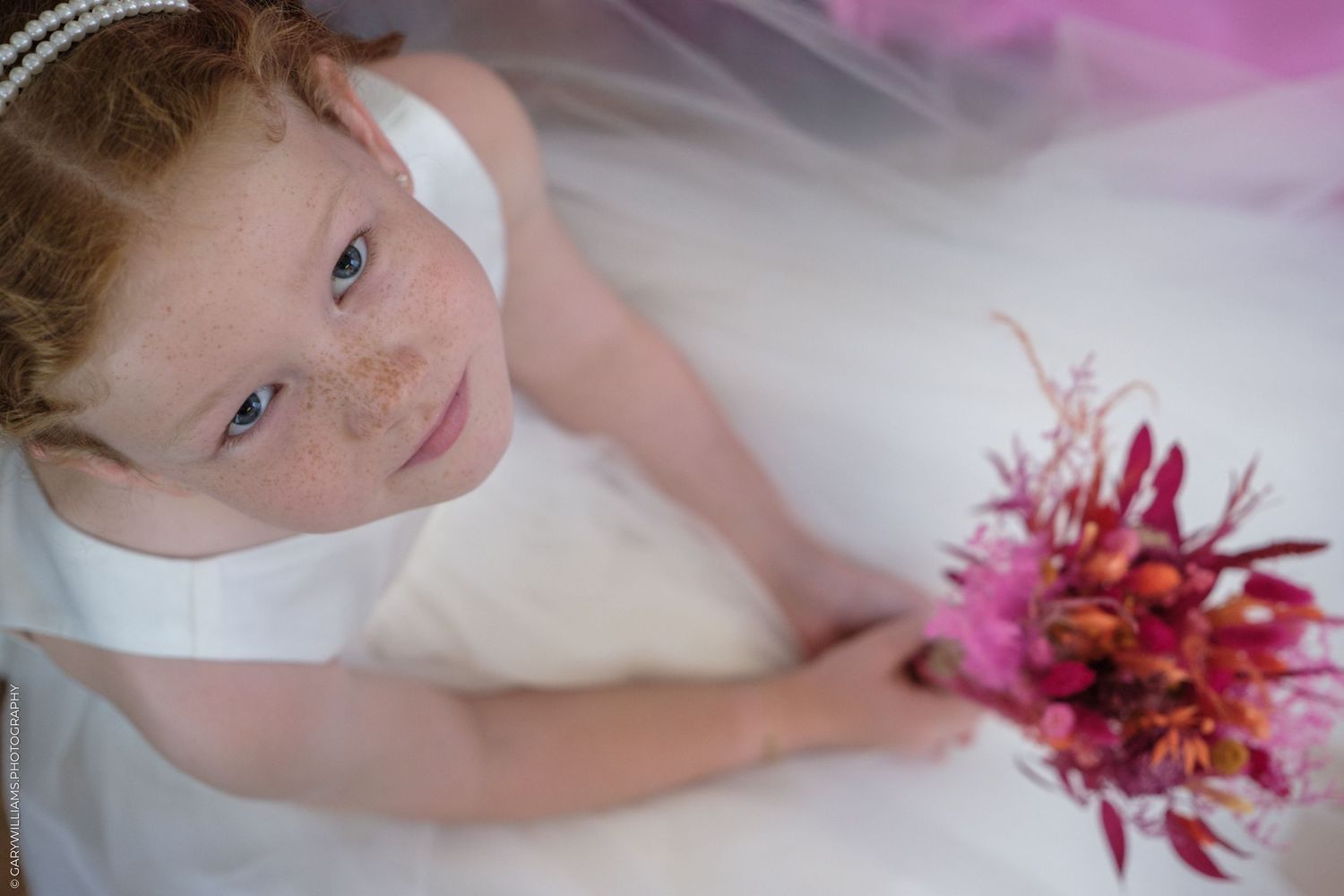 A young flower girl in white dress holding a vibrant pink and red bouquet with a dream-like soft focus background.