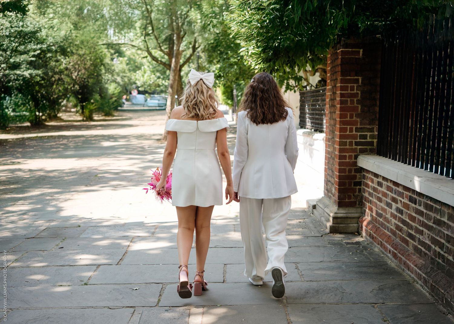 Two people in light-colored outfits walk together along a tree-lined sidewalk next to a brick wall on a sunny day.
