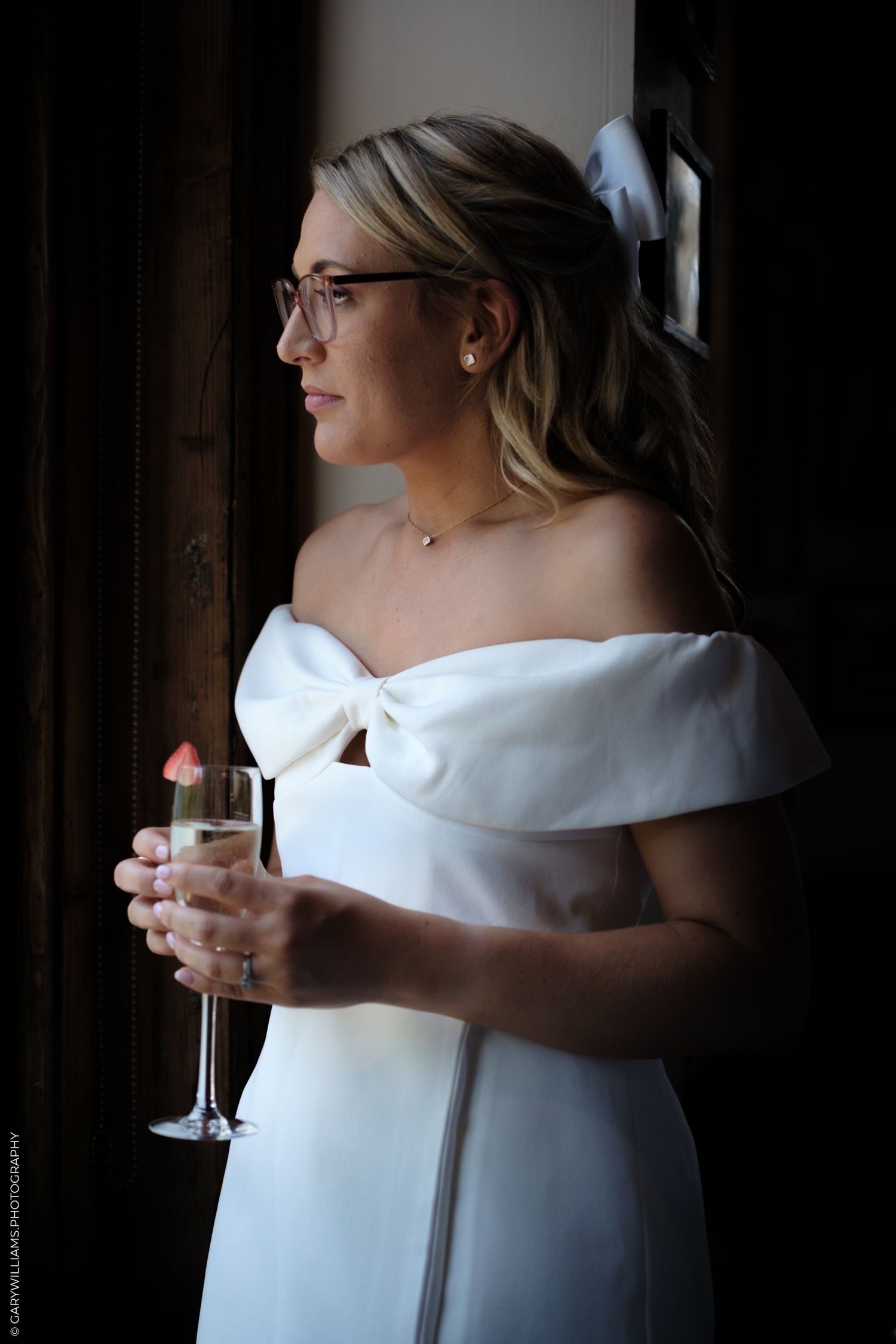 Woman in white off-shoulder dress holding champagne glass and wearing glasses in moody lighting by window.