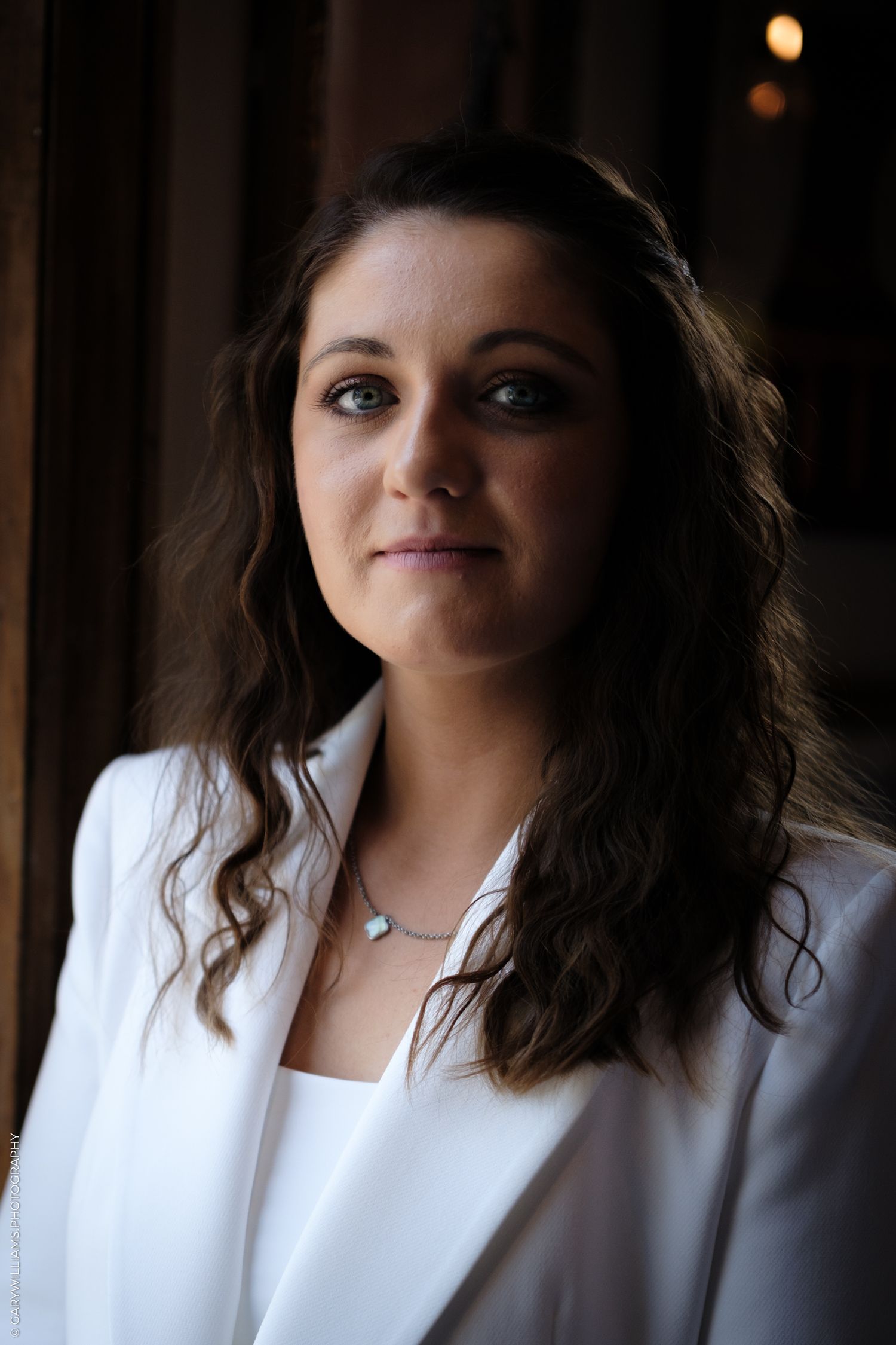 Portrait of a woman in a white blazer and long dark wavy hair against a dark background with soft lighting.
