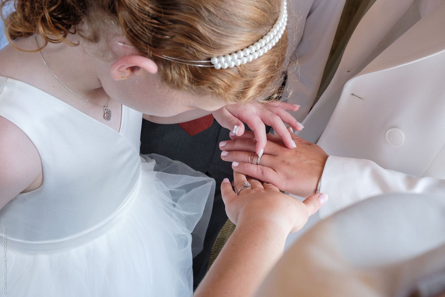 Two hands clasped together wearing wedding attire with a pearl headpiece visible in the background.