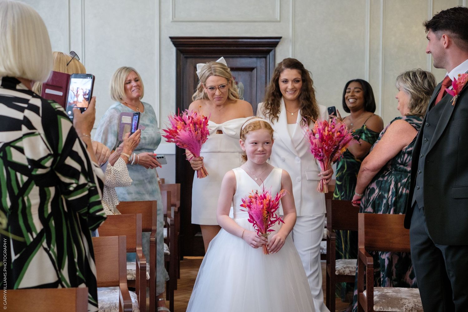 Wedding guests celebrate and capture photos during an indoor ceremony with pink floral bouquets.