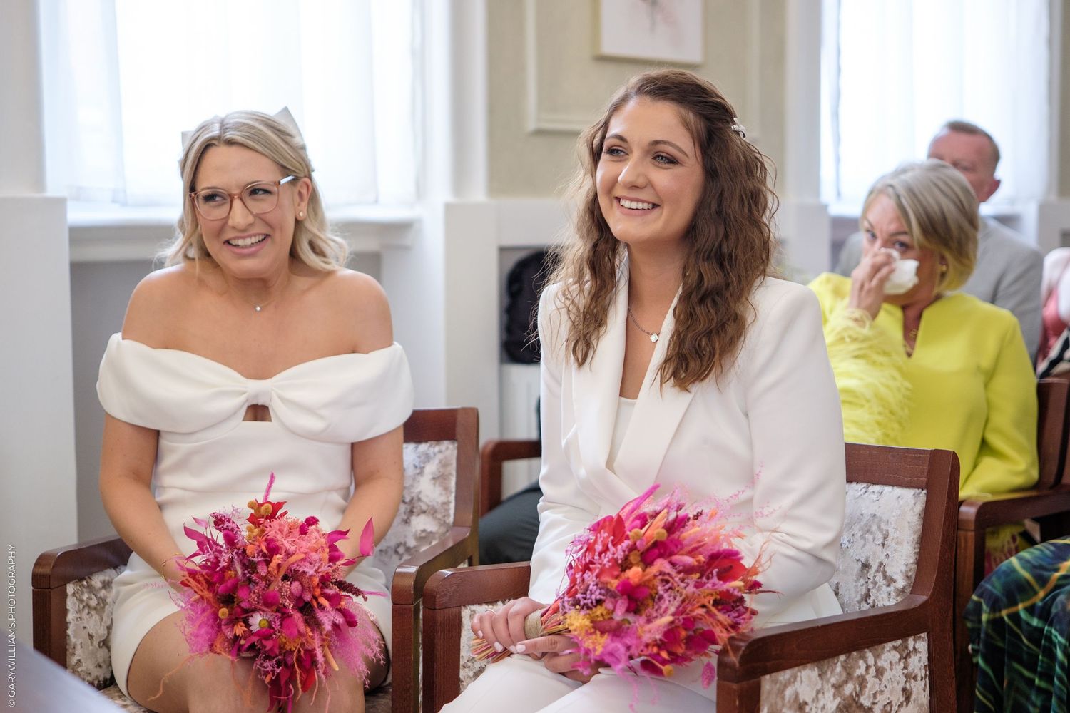 Two people in white dresses hold vibrant pink bouquets while seated and smiling at a special occasion.