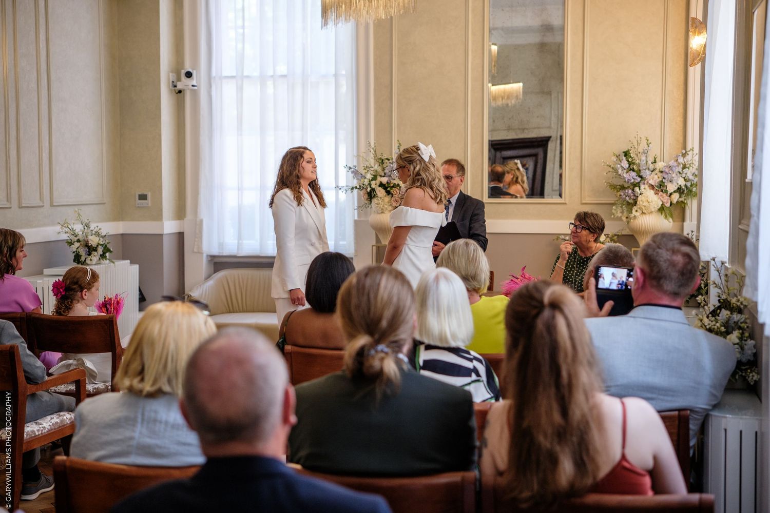 A wedding ceremony taking place in a bright indoor venue with floral arrangements and guests seated in rows.