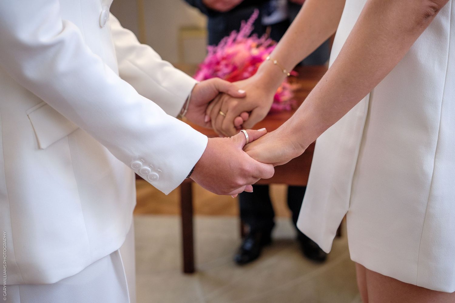 Close-up of hands holding each other during a wedding ceremony, wearing white ceremonial attire.