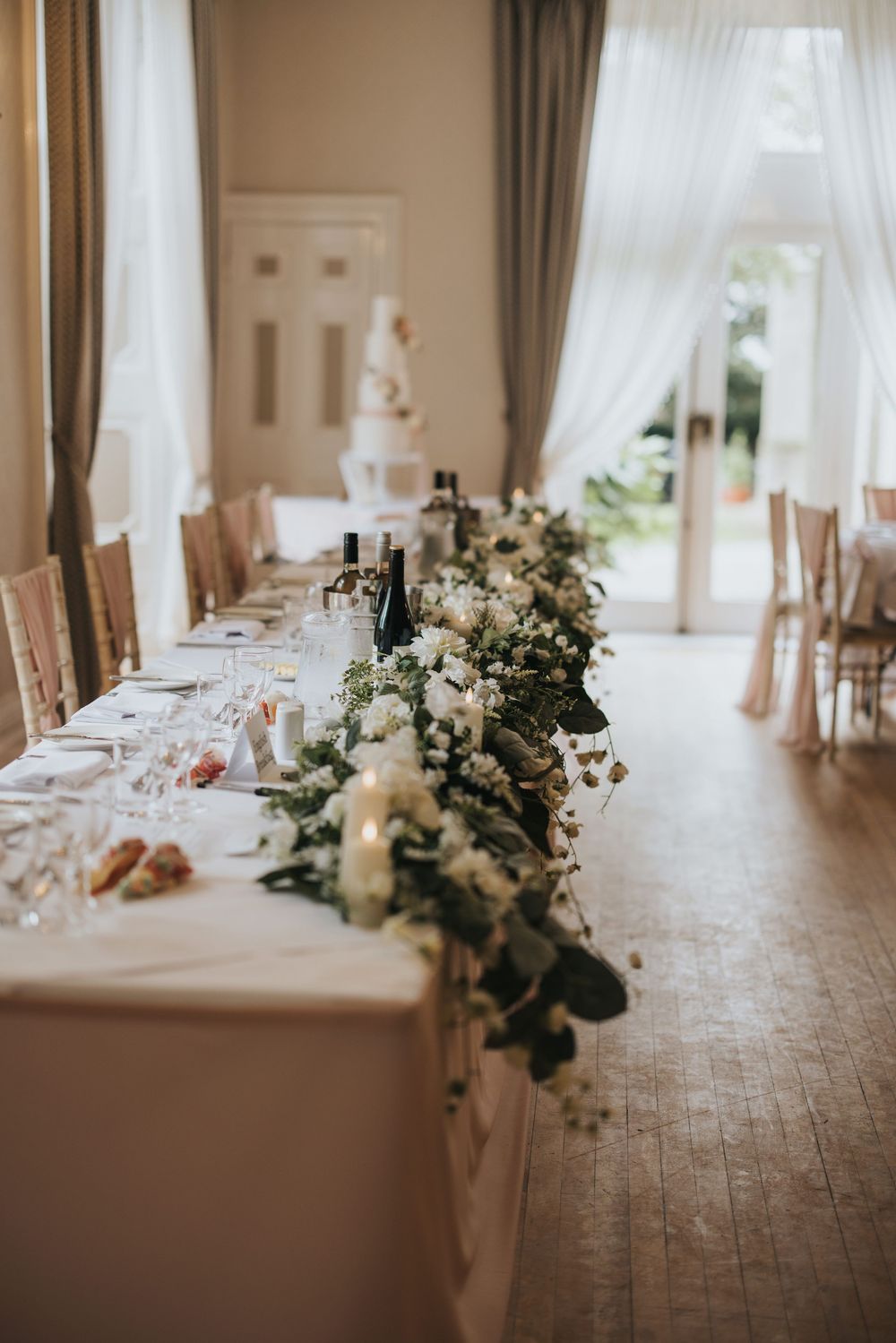 An elegant wedding reception table features a long white floral centrepiece with greenery in a bright sunlit room at Cave Castle.