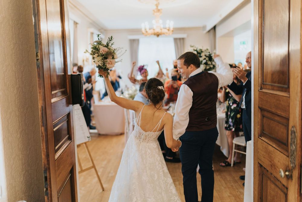 A couple in wedding attire enter a reception venue with their hands raised in celebration as guests cheer at Cave Castle.