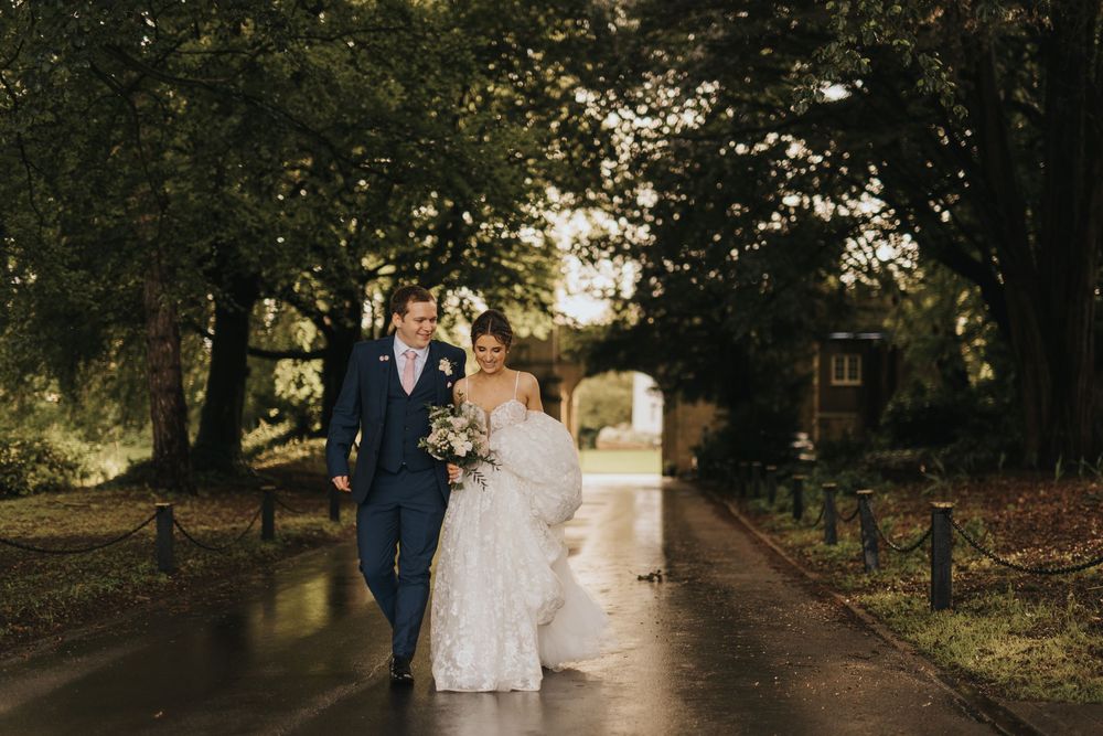 Couple in wedding attire walks down a tree-lined path during autumn as fallen leaves scatter around them at Cave Castle.