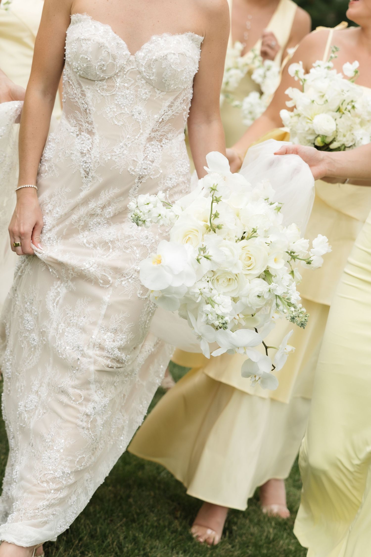 A bride in an ornate lace wedding dress holds a white floral bouquet while bridesmaids in pale yellow dresses stand nearby.