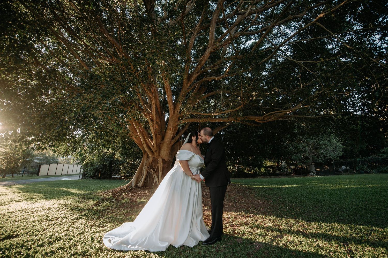 Wedding couple share a romantic embrace under a sprawling tree at sunset during their outdoor ceremony.