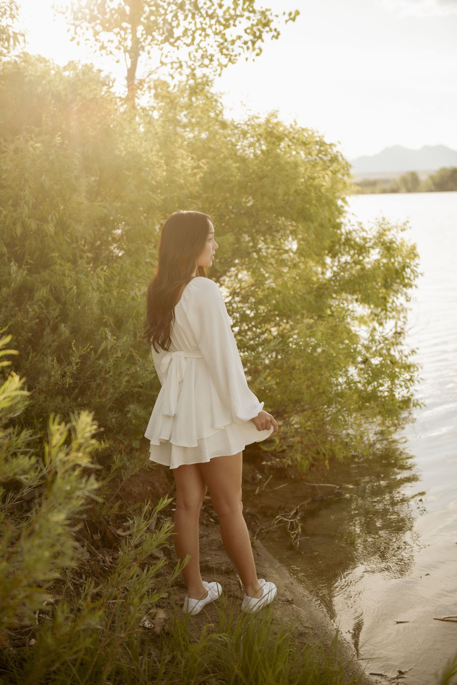 Woman in flowy white dress walking along a serene beach shoreline at sunset with lush greenery nearby.