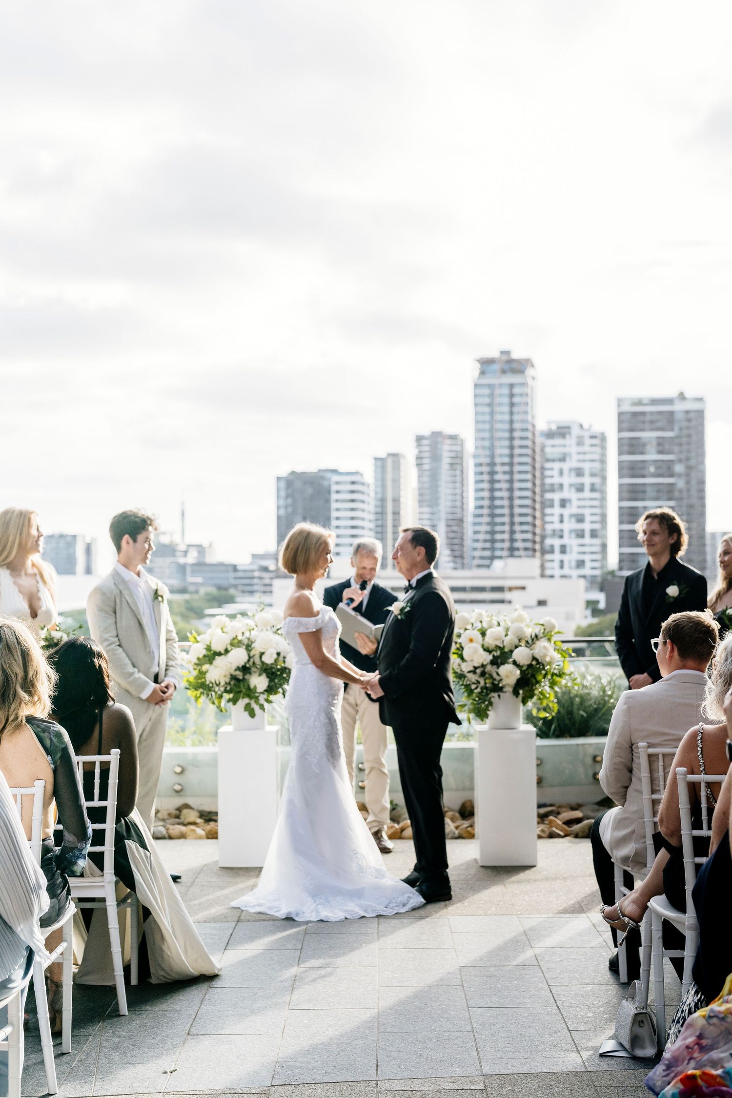 Intimate rooftop wedding ceremony overlooking a modern city skyline with white floral arrangements and white chairs.