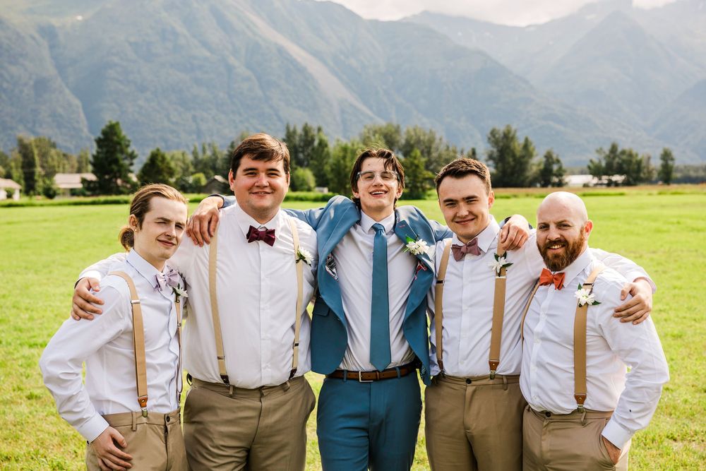 Wedding party members in blue suit and suspenders smile together with mountains in background.