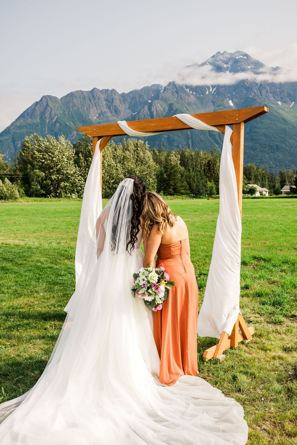 Wedding couple and bridesmaid stand under wooden arch with white draping against stunning mountain backdrop.
