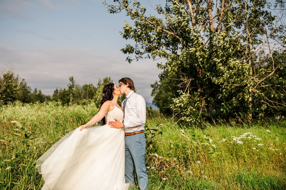 Newlyweds share a romantic moment in a sunlit meadow with mountains in the background.