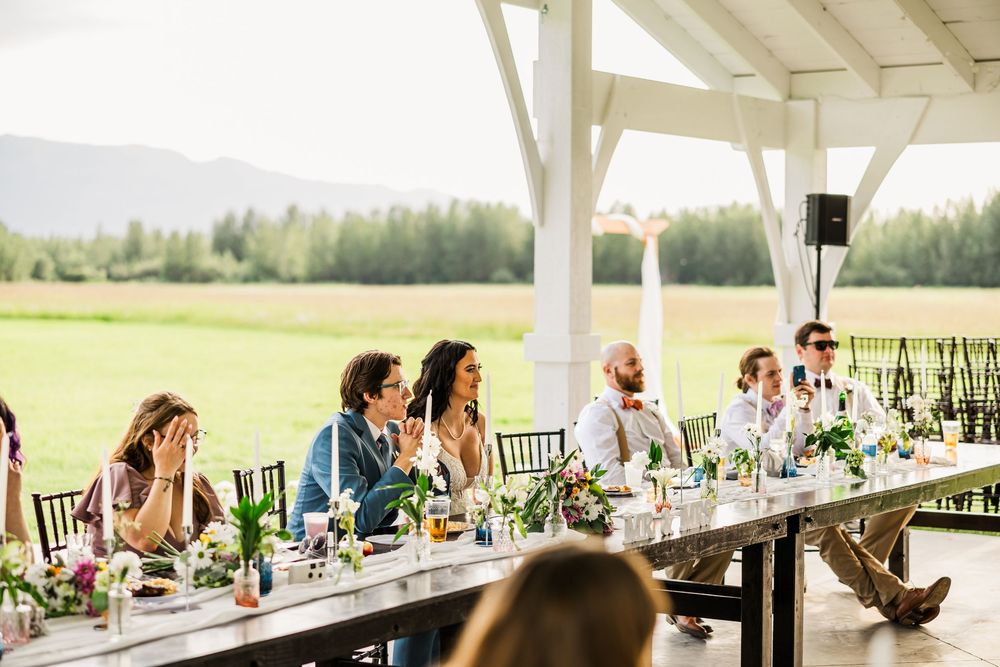 Wedding guests seated at a long outdoor reception table under a white pavilion with mountain views.