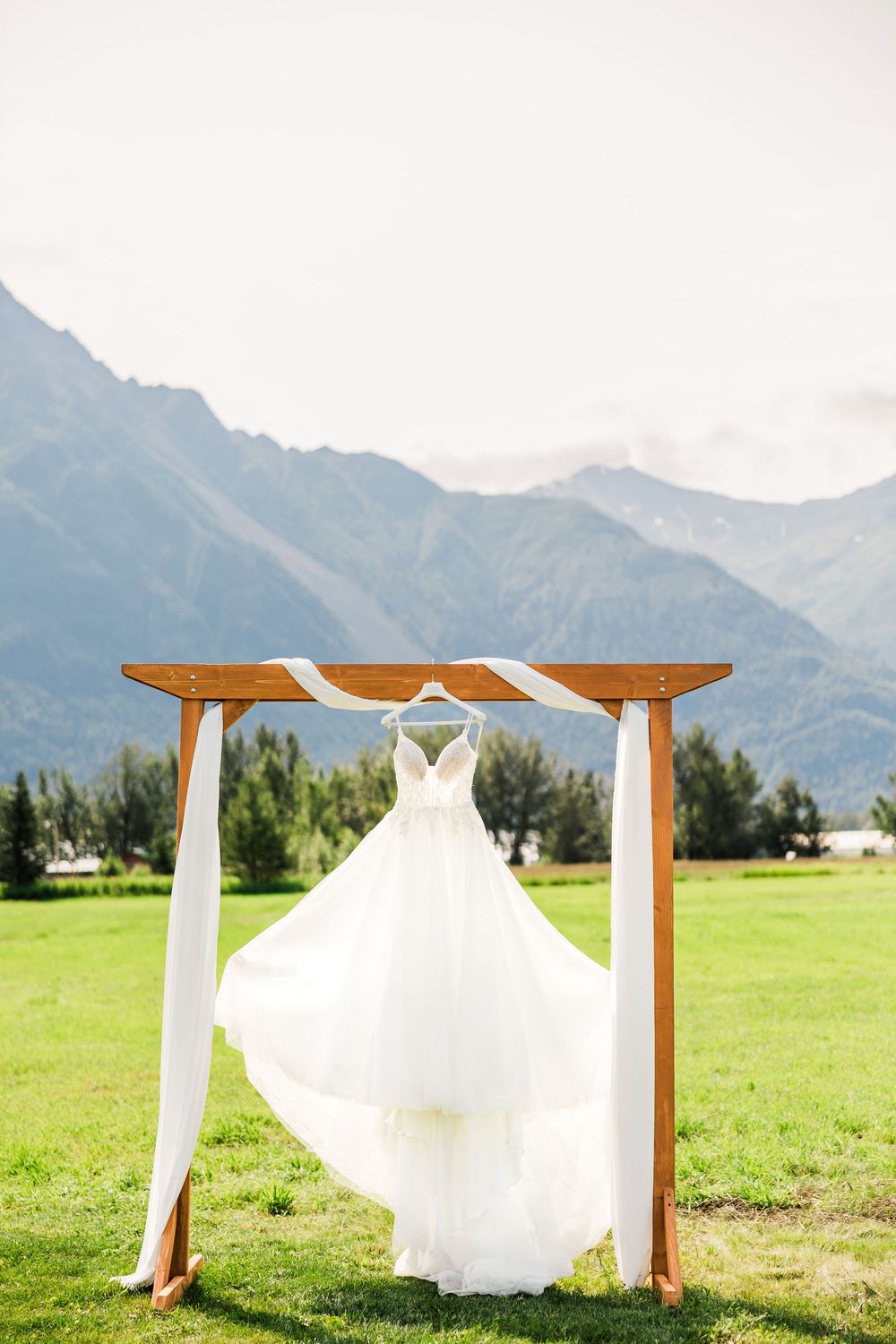 White wedding dress hanging on wooden arch with mountain backdrop.