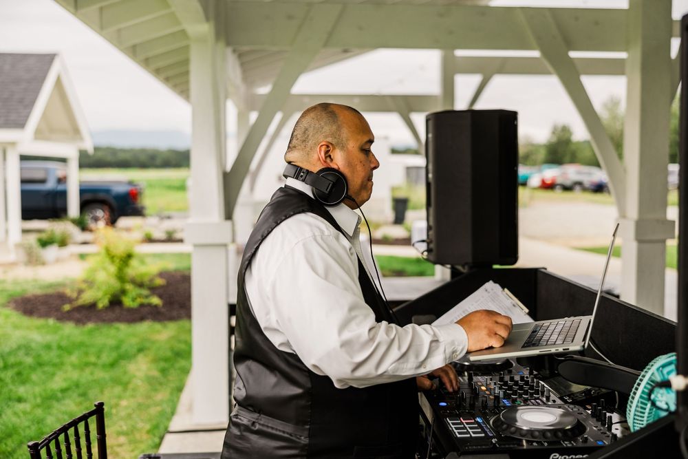 DJ works at laptop computer station under covered outdoor venue during wedding event.