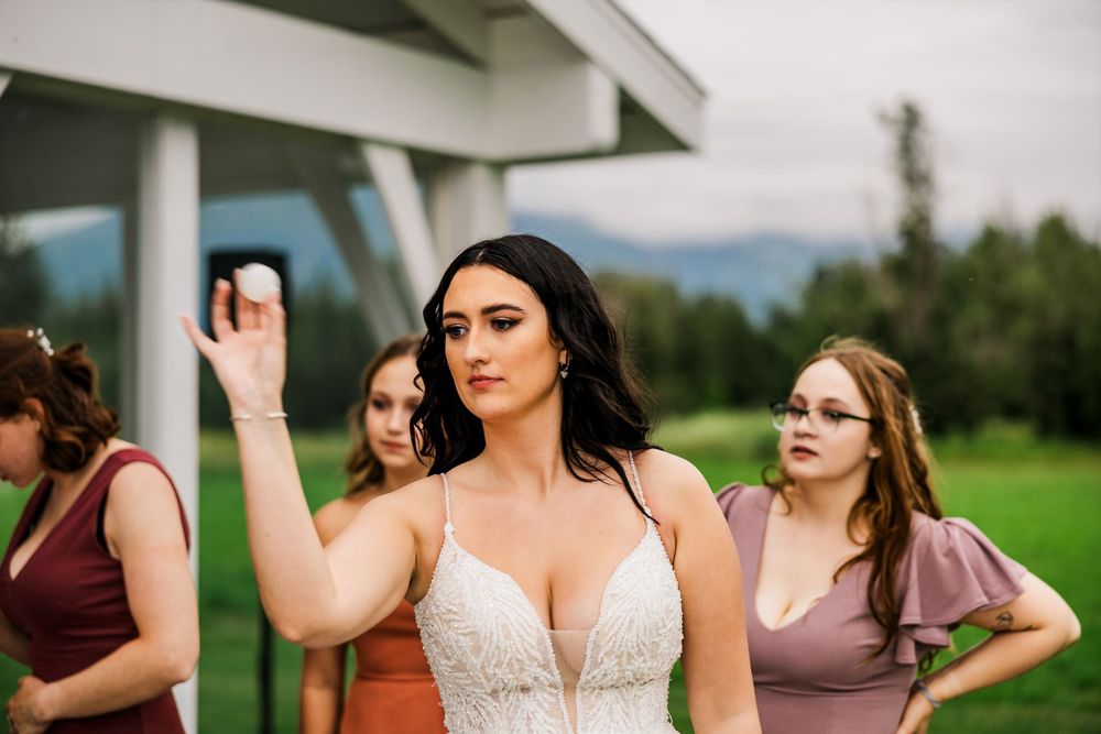 Bride in white dress plays outdoor reception games with bridesmaids in burgundy and sage dresses.