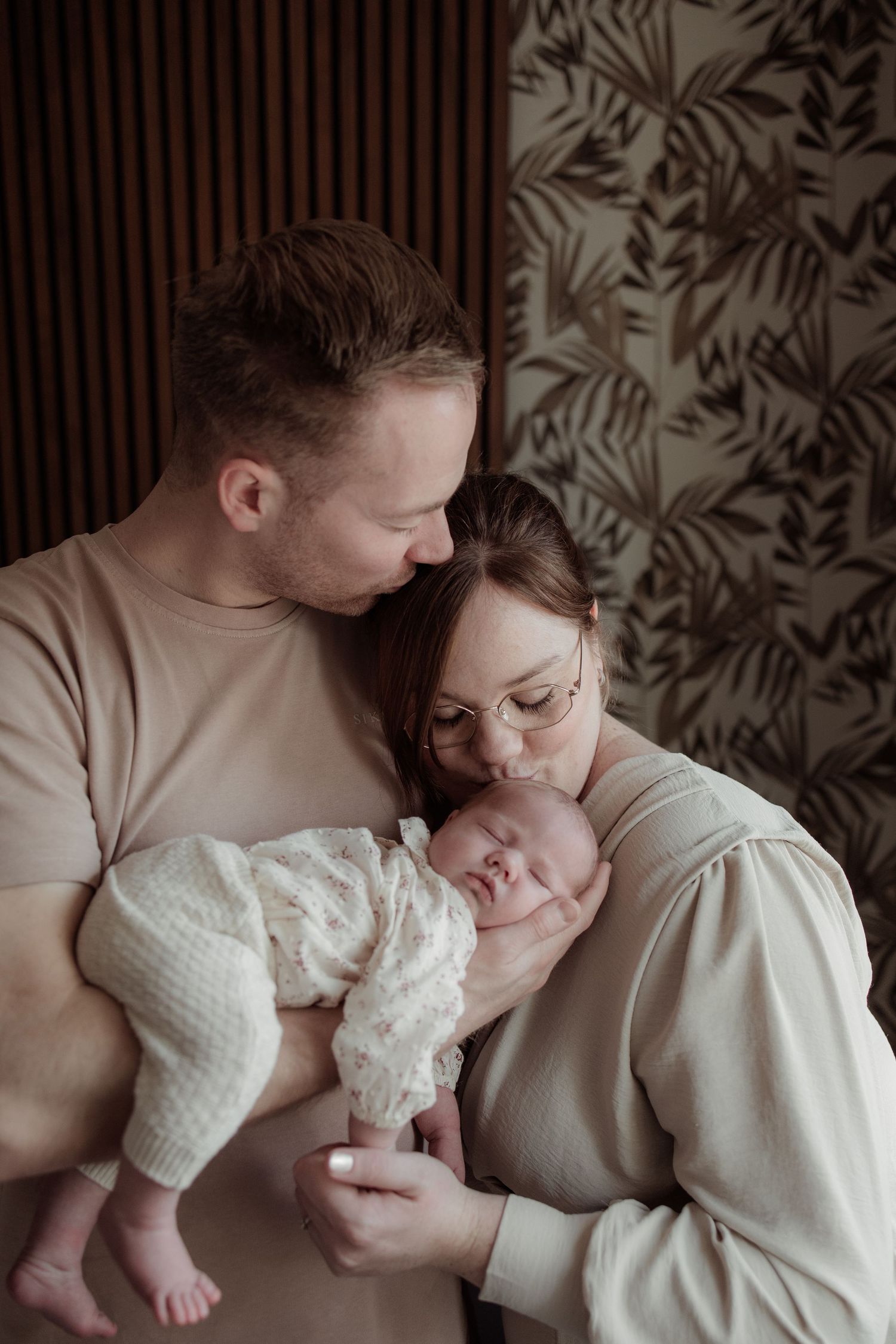 A gentle hand touches a swaddled newborn baby resting on soft beige bedding in moody lighting.