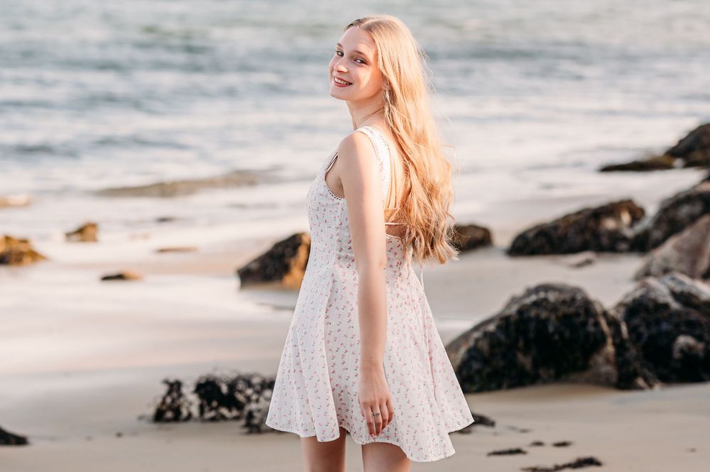 A woman in a white lace dress stands beside ocean rocks on a beach at sunset, facing away from the camera.